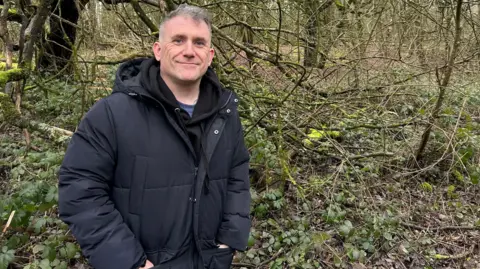 Stew is wearing a dark puffer jacket and has short hair. He is smiling at the camera and standing in the middle of a woodland.