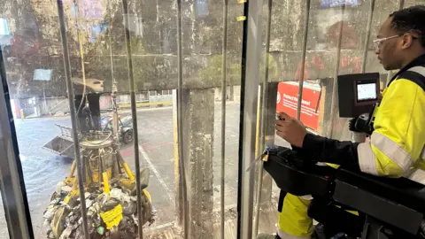 A man wearing glasses and a high visibility outfit is operating machinery which can be seen through a window. It appears to be picking up a pile of rubbish. The man is using a joystick to manoeuvre the machine. 