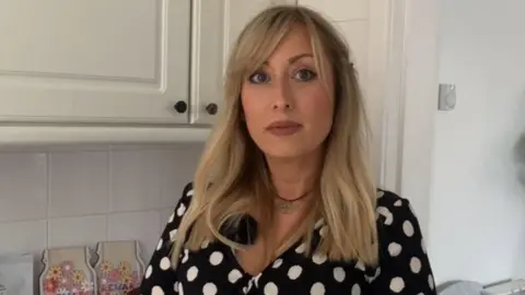 A woman with blond hair, wearing a black and white spotted top looks towards the camera. There are white kitchen cabinets behind her.
