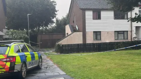 A police car sits beside police tape in front of a house
