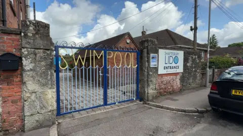 The entrance to the pool site, with an old brick wall and blue gates with the words "Swim Pool" ornately fashioned in the iron, with the shape of waves underneath.