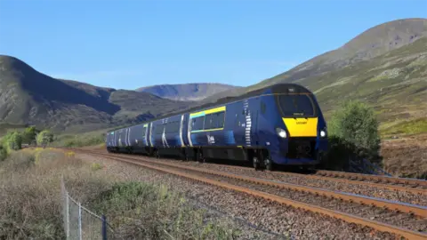 A modern ScotRail train in dark blue livery travels through a scenic Highland landscape, surrounded by mountains, greenery, and clear blue skies.