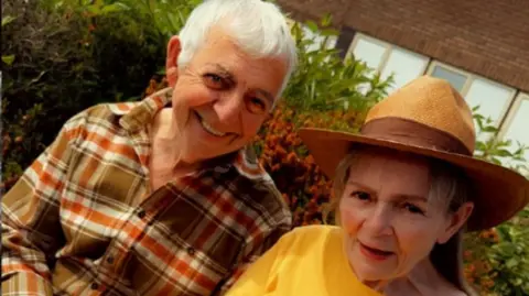 Nicola Hughes Janette with her husband Alex. They are sitting outside. Alex is wearing a checked shirt and Janette is wearing a yellow T-shirt and a hat.