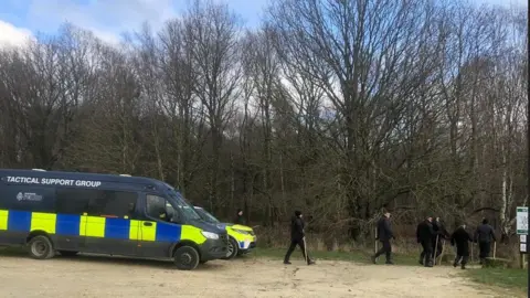 Police officers head into Wombwell Woods to search the woods and lake during, Tactical Support Unit van and police vehicle on the left. Seven police officers dressed in black carrying sticks head into the woods across the centre and right of the picture.