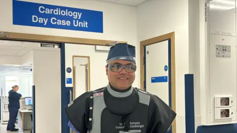 A man dressed in a back gown with plastic glasses and a blue hat standing in a hospital corridor