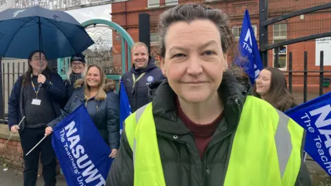 Union rep with black/ grey hair and a high vis jacket stands in front of a school building.