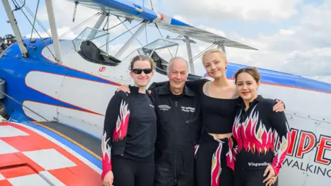 Image of Vic Norman flanked by his three granddaughters Flame Brewer, Rose Powell and Jasmine Norman standing in front of a white blue and red bi-plane