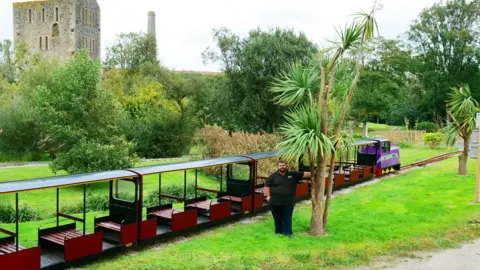 Lappa Valley Ben Harding stood in by one of the newly planted palm trees. The trees are about three times his height. He is stood in the grounds of the tourist attraction, wearing black trousers and a black shirt. He has dark hair and a beard. Directly behind him is one of the trains and it's carriages. Each carriage has red painted wooden seats. The train is a bright purple colour. Behind the train there is grass, bushes a path and an old building which looks similar to a castle. 