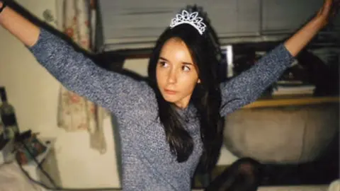 A picture of Fiona Braidwood wearing a long sleeved grey top and a sparkly tiara on her long dark hair. She is sitting in a living room in front of a window, lifting both her arms high and looking off to the left of the camera.