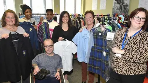 Natalie Frankland stand in a room full of racks of clothes with six other volunteers. They are all smiling broadly at the camera and holding up various pieces of clothing.