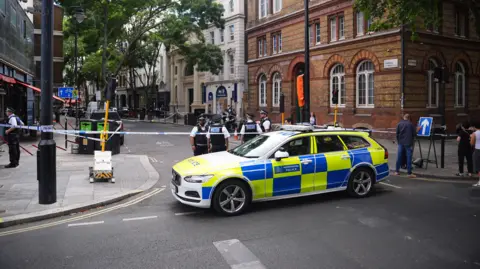 Police car at cordon with officers behind it in front of yellow brick building