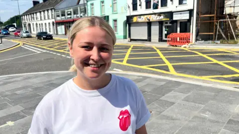 Melissa Caskey, with the box junction behind her. She has blonde hair tied back, wearing a white t-shirt with a red Rolling Stones tongue and lips logo on the left hand side.