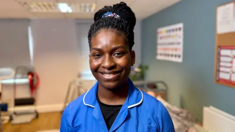 STEVE HUBBARD/BBC Esther Akinpelu stands smiling at the camera. She has braided hair, pulled into a high pony tail. Akinpelu is wearing blue scrubs and is standing in a training room for care staff. There is a bed in the background with a walker and a chart on the wall with the flags of different countries. One wall is green, the other wall is white. The floor is wood laminate.