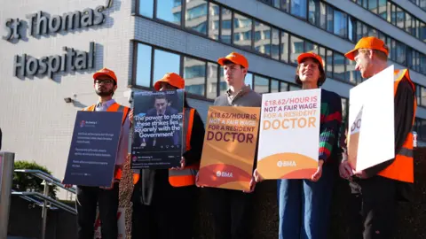 PA Media NHS resident doctors outside St Thomas' Hospital in London on a picket line. They are holding signs and wearing orange hats.