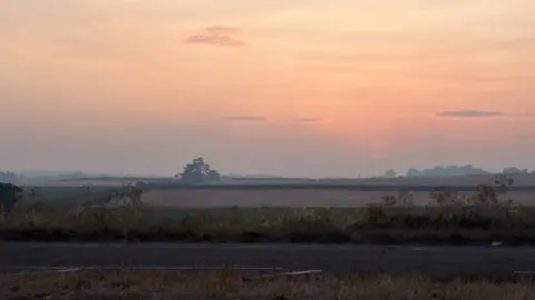 A photograph taken in the early morning shows a clear haze in the air, over rural fields. The sky is orange with the sunrise.