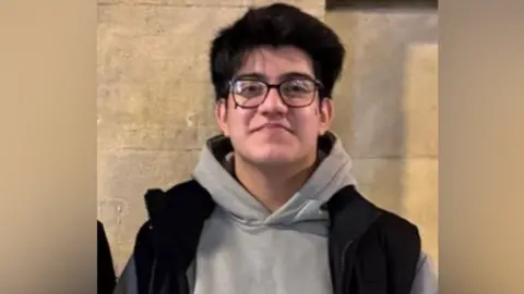 Taha has medium length black her and black rim glasses. He has a grey hoodie and a black gilet 