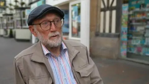 BBC Len Burkitt , who is standing in a street in Stafford wearing a shirt, jacket and cap