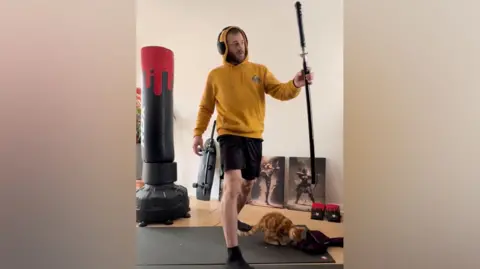 Marcus Arduini Monzo, wearing a mustard yellow hoodie, stands with a samurai sword. His ginger cat sits nearby 