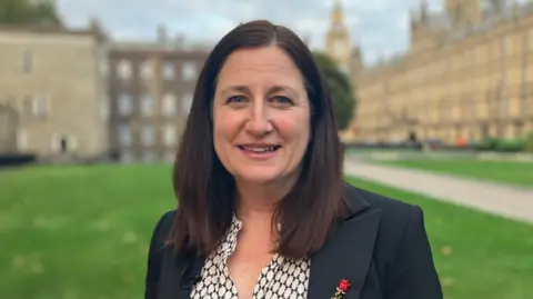 Julia has brown shoulder length hair and is smiling. On her right is the houses of parliament which are large brown buildings. She is wearing a black suit jacket and black and white blouse, with a red rose pin on the jacket