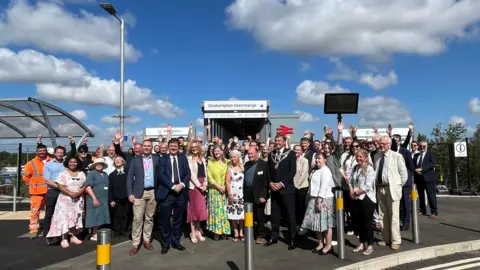 Alison Stephenson A group of people stood at Okehampton station. Some people at the back of the crowd have their hands in the air. 