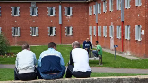 Three male prisoners sit on a wall facing away from the camera and towards the red brick prison building. There is a green courtyard between the inmates and the building.