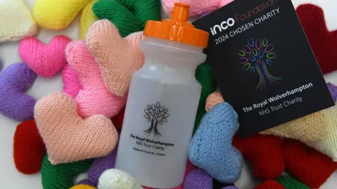 Inco Contracts A white plastic bottle with an orange lid is on top of a pile of multi-coloured knitted hearts. It has a black logo on it that depicts a tree, and says "The Royal Wolverhampton NHS Trust Charity". Next to the bottle is a black leaflet with the same logo on the front.