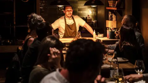 Chef Corrin in kitchen which is open to dining area talking to diners at two tables together. The setting is dark with warm overhead lighting and the diners are all looking at him.