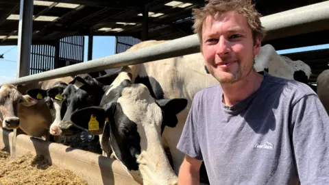 Joe smiles at the camera wearing a blue t-shirt. He crouches down next to some of the cows - which are looking at him curiously, with one sniffing him.