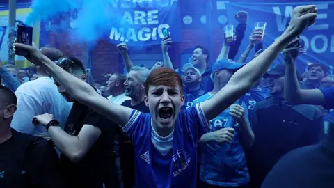Everton supporters outside the stadium with the air thick with blue smoke. A red haired fan has his arms in the air and is shouting