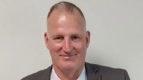 Shropshire Council A short-haired man wearing a dark brown blazer and white shirt smiling in front of a plain, grey background