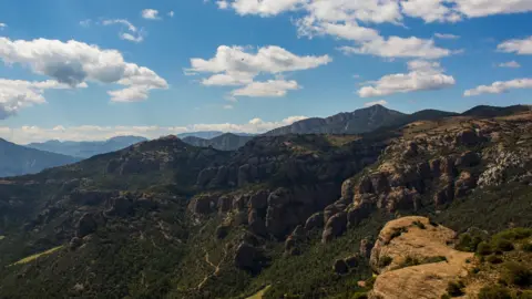 An image depicting the hills of Tremp in Catalonia. Large green and brown mountains stand in front of a blue sky with several clouds.