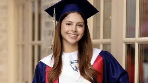 Camila Mendoza Olmos, with long brown hair, smiles and poses for a photo in a red, white and blue graduation cap and gown
