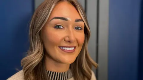 Head and shoulders of Ashley with shoulder-length wavy hair featuring lighter highlights, wearing a light-coloured knit top with a striped neckline. She is looking towards the camera against a softly blurred indoor background with blue wall panels and neutral-toned surfaces.