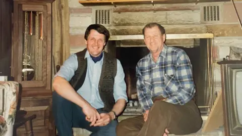 Tim Lobb A younger Tim Lobb is wearing a gilet, a blue shirt and jeans. He is seated next to Marshall Donohew who is in a blue flannelette shirt and brown trousers. They are sitting in front of a fireplace.