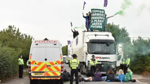 A group of activists blockading a dairy distribution truck. There are several protesters sitting on the floor in front ofa white XPO Logistics truck. There are also several protesters on top of the truck waving flags and green smoke guns. There is a police van next to the truck with several police officers standing amongst the protesters.