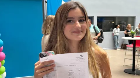 A young woman with light hair holds up her results