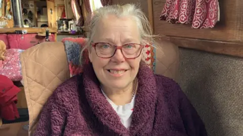 A woman with grey hair, red-framed glasses and a maroon jumper in a brown chair sitting on board her narrowboat. A small room can be seen behind her.
