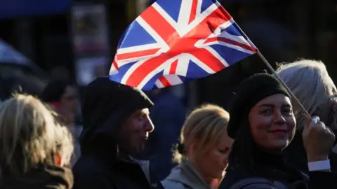 A woman wearing a headscarf,waves a union flag as she stands in a queue. The heads of five other people can also be seen.