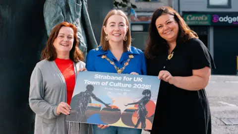 Derry City and Strabane District Council Mayor Grace Uí Niallais is wearing a blue shirt and her mayoral chain. Deirdre Harte is wearing a red shirt and grey cardigan, while Jacqueline Whoriskey is wearing a long black dress. They are all holding the new branding for the Strabane Town of Culture bid.