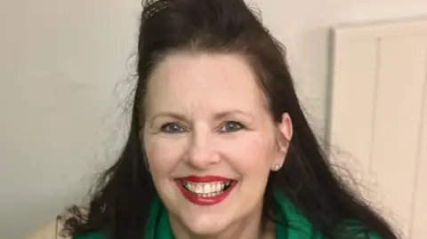 Tanya Wakely Shows woman in long dark hair, an earring in her left ear, red lipstick and a green hoodie smiling