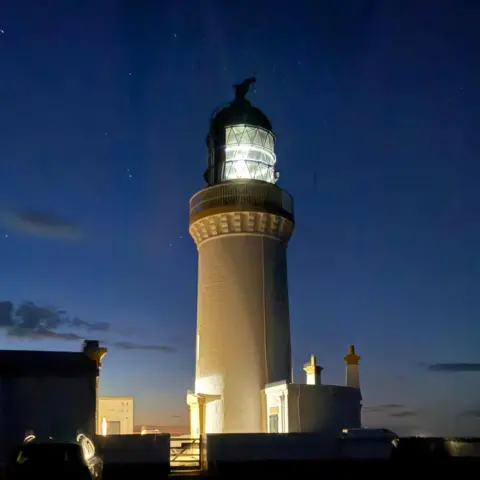 David Fraser A softly lit lighthouse stands against a deep blue night sky, its beacon glowing brightly as the surrounding buildings fade into shadow.