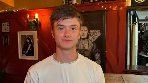 There is a man in a white t-shirt. He has brown hair which is pushed to his right. Behind him is two paintings, one is David Bowie in tudor-inspired clothing. One is a pop star. He is sat next to a wooden table.
