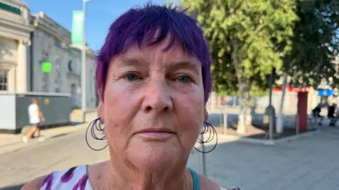 Richard Knights/BBC Mary Williams has short purple hair. She is wearing a white and purple vest top in Clacton town centre and large hoop earrings.
