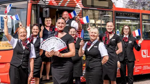 A group of eight women and one man wearing Loganair uniforms smiling at the camera. They are standing in front of a red bus and are waving French flags. The steward at the front is holding a bunch of flight tickets fanned out. The unform is black and the women are wearing red checked scarfs.