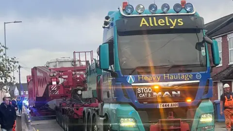 A truck pulling a large red piece of machinery down Tomline Road in Ipswich. There are houses to the right of the truck. There are workers in high visibility overalls on each side and cones on the left hand side of the road.
