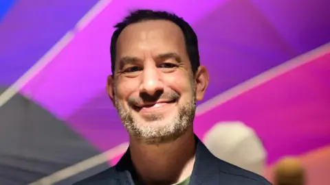 Jonathan Harper smiling at the camera. He has dark hair worn in a short style and has a slightly grey-ing beard.  He is wearing a green T-shirt and a charcoal or navy blazer. Harper is standing in front of a rainbow hot air balloon.
