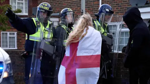 PA Officers with riot shields talk to a woman draped in a St George flag and a man wearing a dark hoodie. 