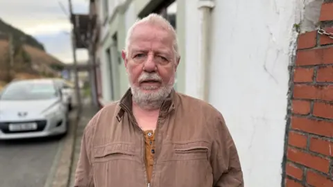 An old man with a stern expression looking towards the camera. He has grey hair, beard and moustache. He is wearing a brown coat with a lighter brown top underneath. His background consists of a brick wall to his right and some parked cars to his left.