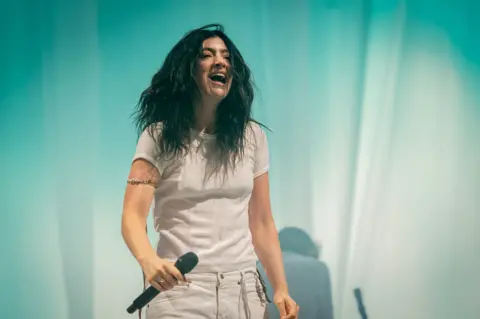 Singer Lorde wears a white t-shirt and trousers in front of a turquoise curtain on-stage. She's holding a microphone and smiling.