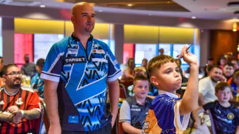 Oscar has short brown hair, he is wearing a blue and yellow sports top throwing a dart. Darts player Rob Cross is stood behind him - he is bald and wearing a blue sports top. A crowd of people watch in the background. 